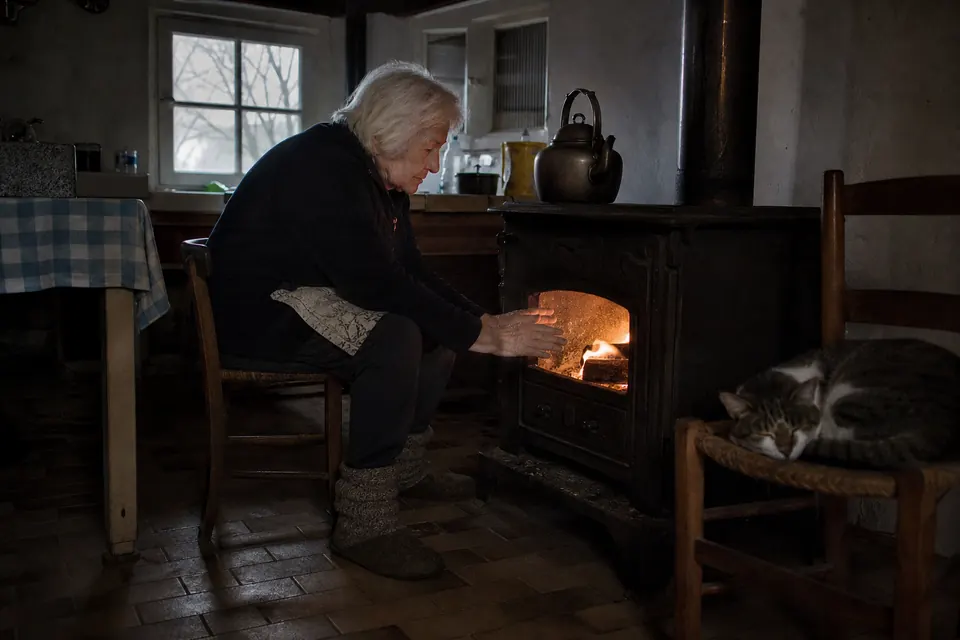 Femme âgée se réchauffant les mains devant un poêle à bois dans une cuisine de ferme