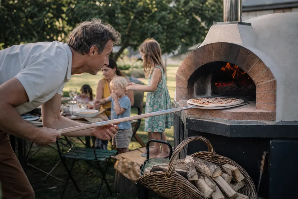 Famille autour d'un four à pizza au bois dans un jardin, un homme enfourne une pizza