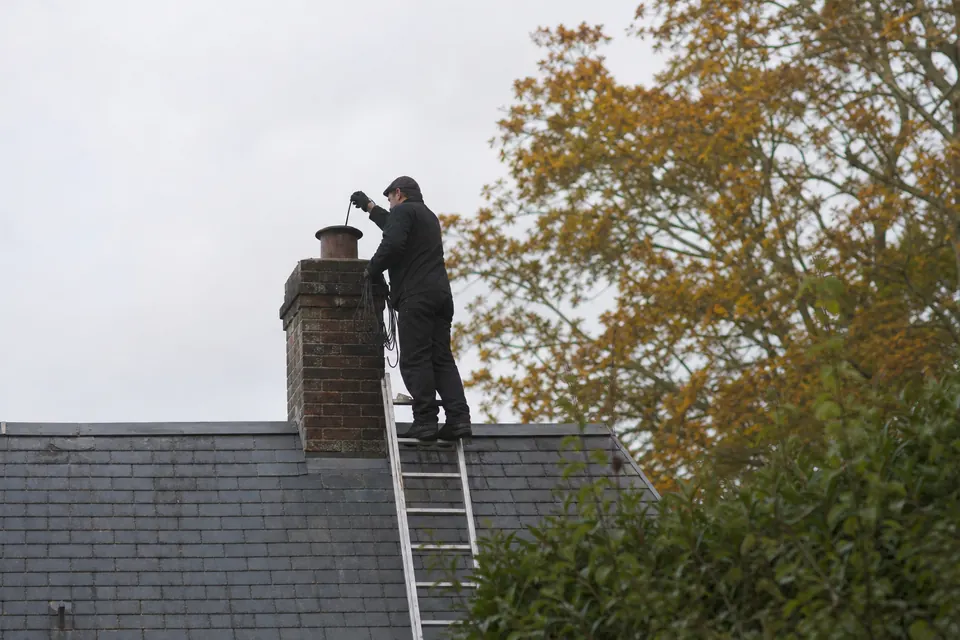 Ramoneur sur une échelle nettoyant la cheminée d'une maison normande au toit d'ardoise