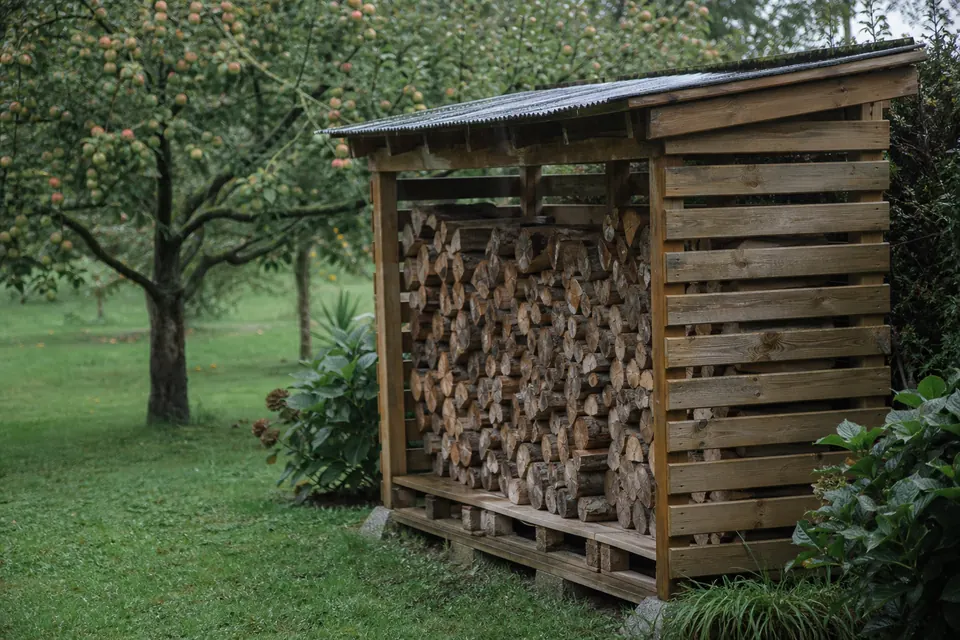 Abri à bois ventilé avec bûches rangées sur palettes dans un jardin normand sous la bruine