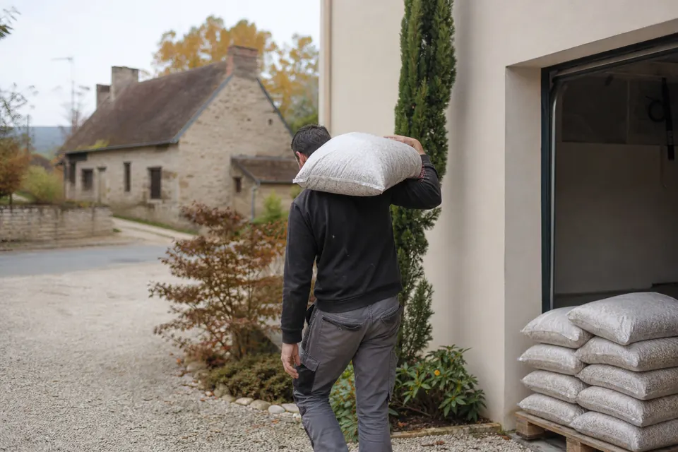 Homme portant un sac de granulés sur l'épaule vers un garage dans un village