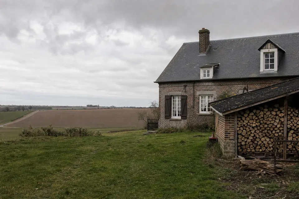 Plaine agricole normande avec une ferme en briques et une réserve de bois sous un appentis